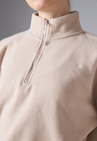 Pulover din fleece de un bej moale, cu guler înalt și închidere cu fermoar pe jumătate. Prezintă un logo subtil pe piept și un material texturat.