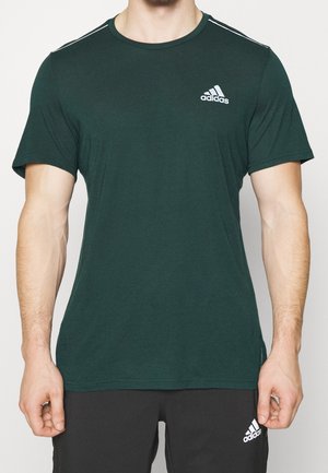 Hombre con camiseta Adidas verde oscuro y pantalones cortos negros Adidas, de pie con los brazos relajados a los lados contra un fondo liso.