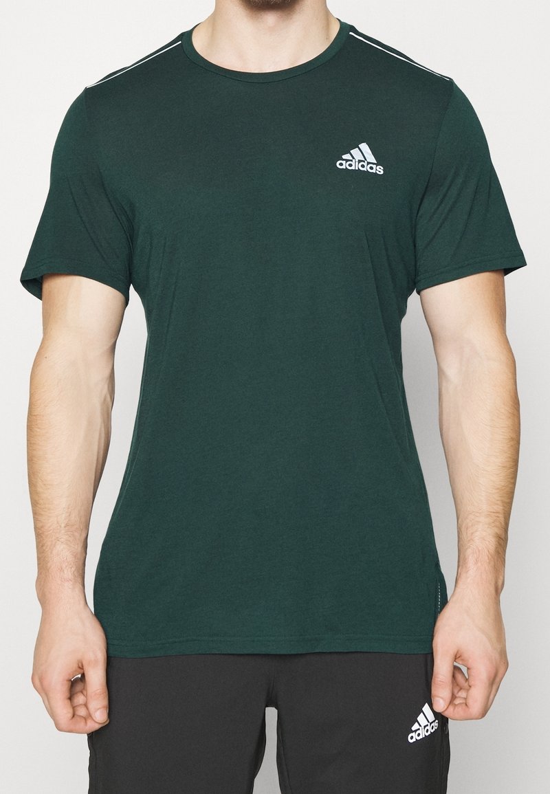 Homme portant un T-shirt Adidas vert foncé et un short Adidas noir, debout les bras détendus le long du corps devant un fond uni.