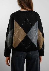 Persona che indossa un maglione nero con grandi motivi a diamante marroni e grigi, abbinato a pantaloni scuri, mostrato di spalle contro uno sfondo neutro.