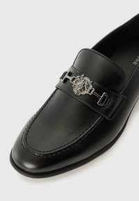 Mocasín de piel negra con un acabado liso, que presenta un acento de emblema de león y herrajes plateados, además de una costura detallada a lo largo del borde.