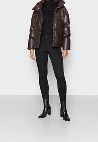 Brązowa kurtka puchowa z wysokim kołnierzem, zestawiona z czarnymi obcisłymi jeansami i czarnymi botkami na obcasie. Gładka, błyszcząca faktura, minimalistyczny design.