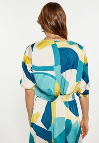 Donna con i lunghi capelli castani che indossa un abito con stampa astratta multicolore, caratterizzato da forme turchesi, gialle e bianche, vista da dietro.