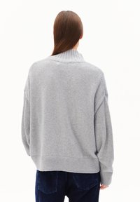 Personne avec de longs cheveux bruns portant un pull léger gris clair et un pantalon en velours côtelé bleu foncé, montré de dos sur un fond blanc.