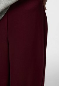 Bordowe spodnie wykonane z gładkiej tkaniny o szerokich nogawkach. Posiadają boczny szew i subtelny układ fałd dla swobodnego kroju.
