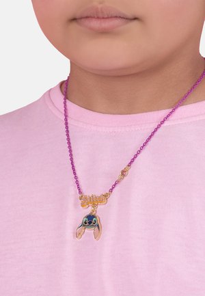 Collier de perles violettes avec une étiquette en or "Stitch" et un charm en forme de Stitch coloré avec des oreilles proéminentes, le tout sur fond de chemise rose.