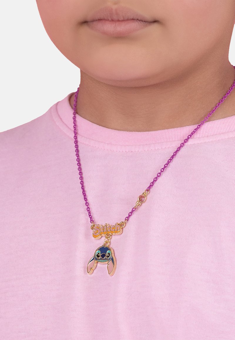 Collier de perles violettes avec une étiquette en or "Stitch" et un charm en forme de Stitch coloré avec des oreilles proéminentes, le tout sur fond de chemise rose.