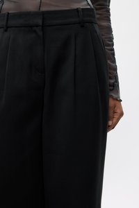 Black tailored trousers with front pleats, belt loops, and a smooth texture. A hand is visible resting on the side seam.
