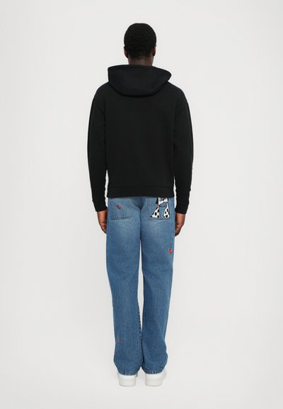 Felpa nera con cappuccio e vestibilità comoda, abbinata a jeans blu larghi con un motivo di un cane ricamato e piccoli dettagli rossi. Scarpe da ginnastica bianche indossate.