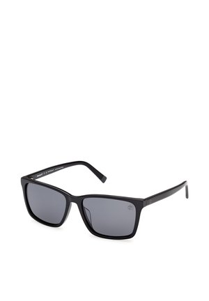 Schwarze rechteckige Sonnenbrille mit dunkel getönten Gläsern und schlanken Bügeln, aus einem frontalen Seitenwinkel auf weißem Hintergrund gezeigt.