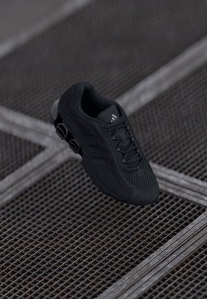Sort Adidas løbesko med fjederbelastet sål, ophængt over metalkryds.