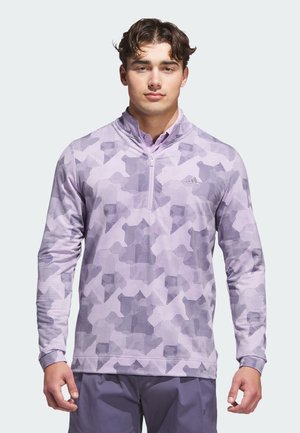 Giovane uomo che indossa un pullover a mezza zip di colore viola chiaro con motivo a quadretti e pantaloni viola scuro, in piedi davanti a uno sfondo chiaro e uniforme.