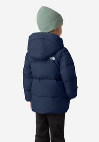 Námořnický puffer kabát s kapucí, který má prošívanou texturu a logo na levém rameni, nošený s světle modrým pleteným čepicím.