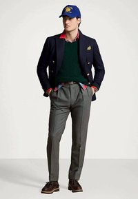 Un homme en blazer marine, pull vert, chemise à carreaux rouges, pantalon gris plissé, chaussures marron et casquette bleue, se tenant les mains dans les poches.