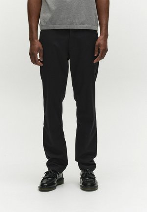 Mees, kes kannab musti sirge säärega pükse, musti narmastega mokkasaapaid ja halli t-särki, seisab valge tausta ees.