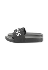 Fekete slip-on szandálok texturált talpbéléssel és széles pántokkal, melyeken nagy fehér betűk félig látszanak egy fehér háttéren.