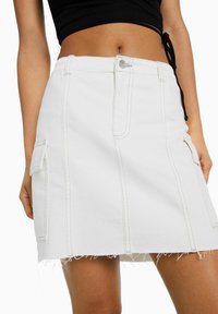 Bílá riflová sukně s třepícím se lemem, která má dvě postranní kapsy, kontrastní švy a zapínání na knoflík v pase.