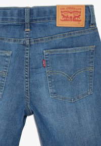Jeans de mezclilla azul con bolsillos traseros visibles, costuras doradas y una etiqueta de cuero marrón en la cintura con el logotipo de la marca y la información de tamaño.