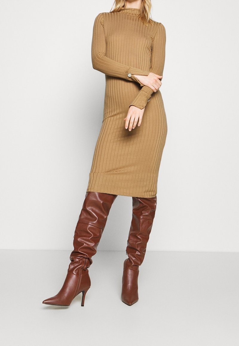 Femme portant une robe midi beige cintrée à manches longues côtelées, associée à des bottes hautes en cuir marron souple à bout pointu et talons.