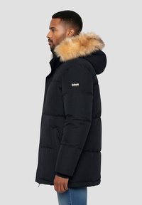 Abrigo acolchado negro con capucha forrada de piel, con cierre de cremallera y bolsillos laterales. El diseño presenta una textura lisa y un patrón acolchado rectangular.