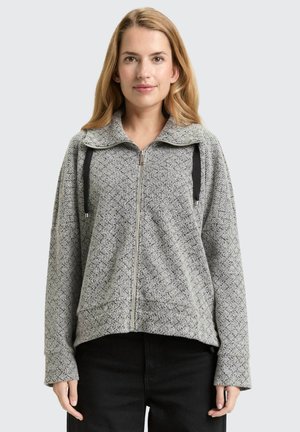 Femme portant un sweat à capuche gris à fermeture éclair avec un col montant et des cordons noirs, associé à un pantalon noir, faisant face à l'avant sur un fond clair.