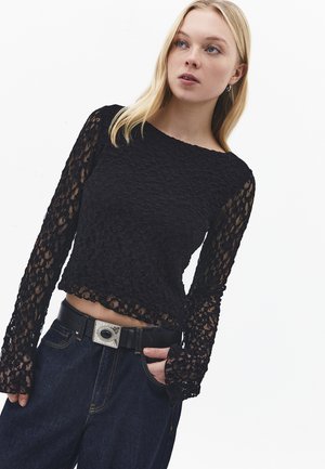 Jeune femme aux cheveux blonds porte un haut en dentelle noire à manches longues et un jean bleu foncé avec une ceinture noire, posant devant un fond uni.