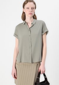 Short-sleeve, collared shirt in light olive green, featuring a button front and soft fabric. Model wears beige pinstripe skirt and holds a black bag.
