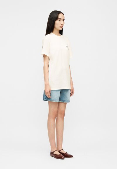 Woman standing facing forward wearing a plain off-white T-shirt, light blue denim shorts, and brown Mary Jane flats.