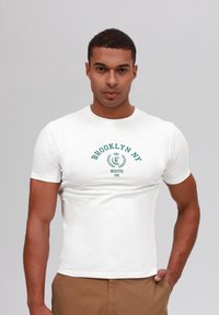 Camiseta blanca de manga corta con el texto impreso en verde "BROOKLYN NY" con acentos de laurel, diseño ajustado y textura de algodón suave.