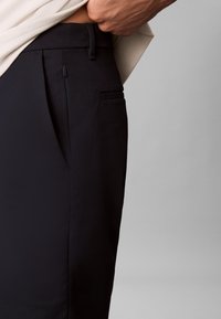 Pantaloni negri cu textură netedă, prevăzuți cu buzunar lateral, curele pentru centură și o croială elegantă, parțial acoperiți de o cămașă deschisă la culoare.