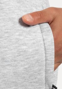 Lichtgrijze sweatshirtstof met een zachte textuur; heeft een zijzak en zichtbare stikseldetails. Hand rustend op de opening van de zak.