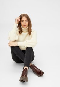 Suéter de cuello alto de canalé crema con mangas voluminosas, combinado con jeans oscuros ajustados y botines marrones. Fondo neutro.