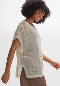 Pull en maille ouverte beige avec des manches courtes, un col rond et des fentes sur les côtés. Sa texture légère favorise la respirabilité et le confort.