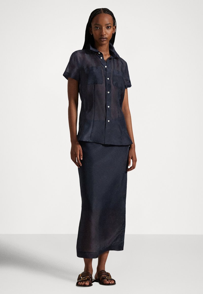 Frau trägt eine dunkle, halbtransparente Kurzarm-Bluse mit Knöpfen und einen passenden langen Rock sowie braune Sandalen, steht vor einem einfarbigen Hintergrund.