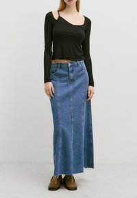 Schwarzes Langarmoberteil mit asymmetrischem Ausschnitt, kombiniert mit einem blauen Jeans-Maxikleid mit fransigem Saum und vorderen Nahtdetails. Braune Stiefel.