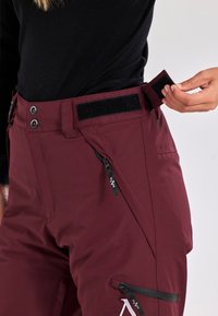 Pantaloni da neve color bordeaux con una cintura in velcro, due tasche laterali con cerniere e un piccolo logo sul lato anteriore destro in basso.
