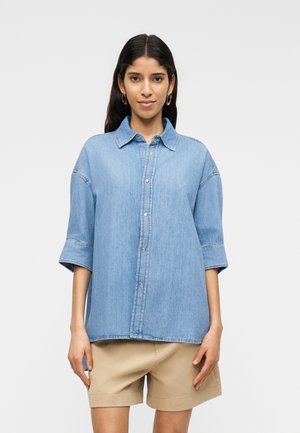 Nainen, jolla on pitkät tummat hiukset, kullanväriset rengaskorvakorut, vaaleansininen farkkupaita, jossa on käärityt hihat, ja beiget shortsit.