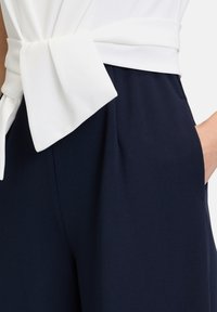 Pantalons navy avec une texture lisse, dotés d'une ceinture en tissu blanc à la taille, complétés par des poches latérales et de légères plis.