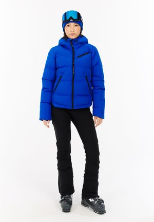 Blauwe geïsoleerde jas met een capuchon, ritsen en zwarte accenten, gecombineerd met zwarte skibroeken en zwarte skischoenen. Beschikt over een strak, compact ontwerp.