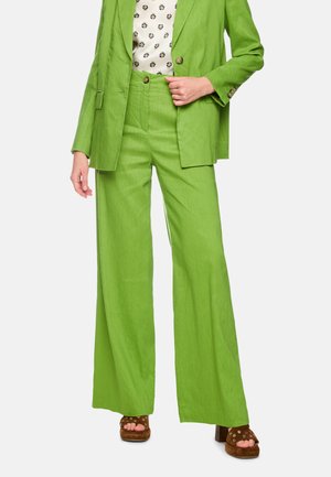 Woman wearing bright green wide-leg trousers and matching blazer, paired with patterned blouse and brown heeled sandals.