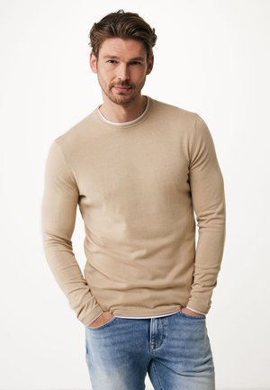 Beige langslavige trui met een ronde hals, geribde manchetten en een gladde textuur, gedragen over lichtblauwe spijkerbroeken.