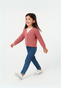 Rosa ribbatten kofta med tre knappar, bärs över en vit topp. I kombination med blå jeans och vita sneakers. Barn som går på en ljus bakgrund.