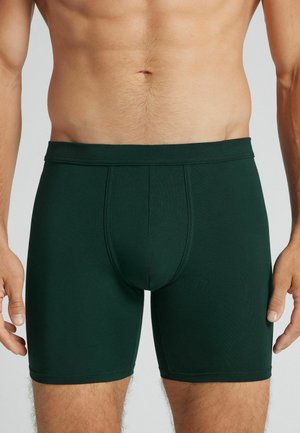 Torse masculin portant un boxer vert foncé, debout avec les bras détendus le long du corps sur un fond neutre.