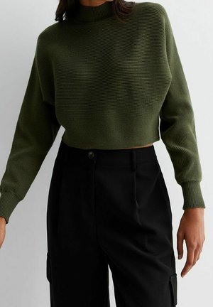 Personne portant un pull court côtelé vert olive et un pantalon noir taille haute, vue des épaules aux cuisses sur un fond uni.