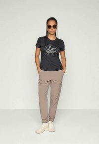 Camiseta negra de manga corta con un gráfico circular, combinada con pantalones jogger beige claros y zapatillas beige. Unas gafas de sol redondas completan el look.