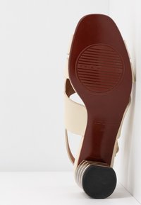 Vue de la semelle d'une sandale à talon beige avec semelle marron, nom de marque embossé et talon rond noir empilé, positionnée contre un coin blanc.