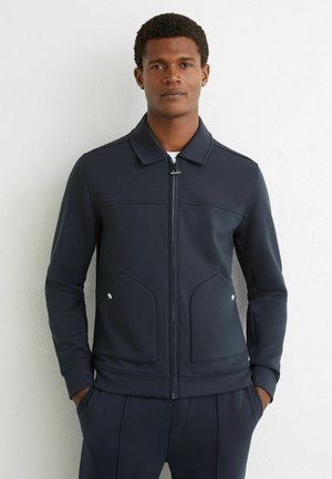 Mann trägt eine marineblaue Jacke mit Reißverschluss, Vorderklappentaschen und passende marineblaue Hose, steht vor einer weißen Wand.