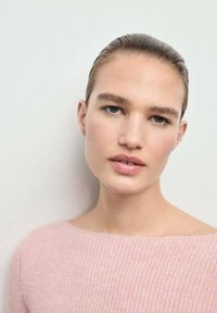 Giovane con capelli all'indietro, che indossa un maglione rosa pallido, guarda dolcemente la fotocamera su uno sfondo chiaro e semplice.