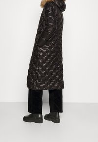 Abrigo de plumas negro largo y acolchado con capucha y borde de piel. Presenta bolsillos laterales y una textura brillante. Lleva puesto pantalones negros cortos y botas gruesas.