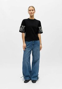 Femme debout portant un haut noir à manches courtes avec des broderies blanches sur les manches et un jean bleu à jambes larges avec des liens aux ourlets.